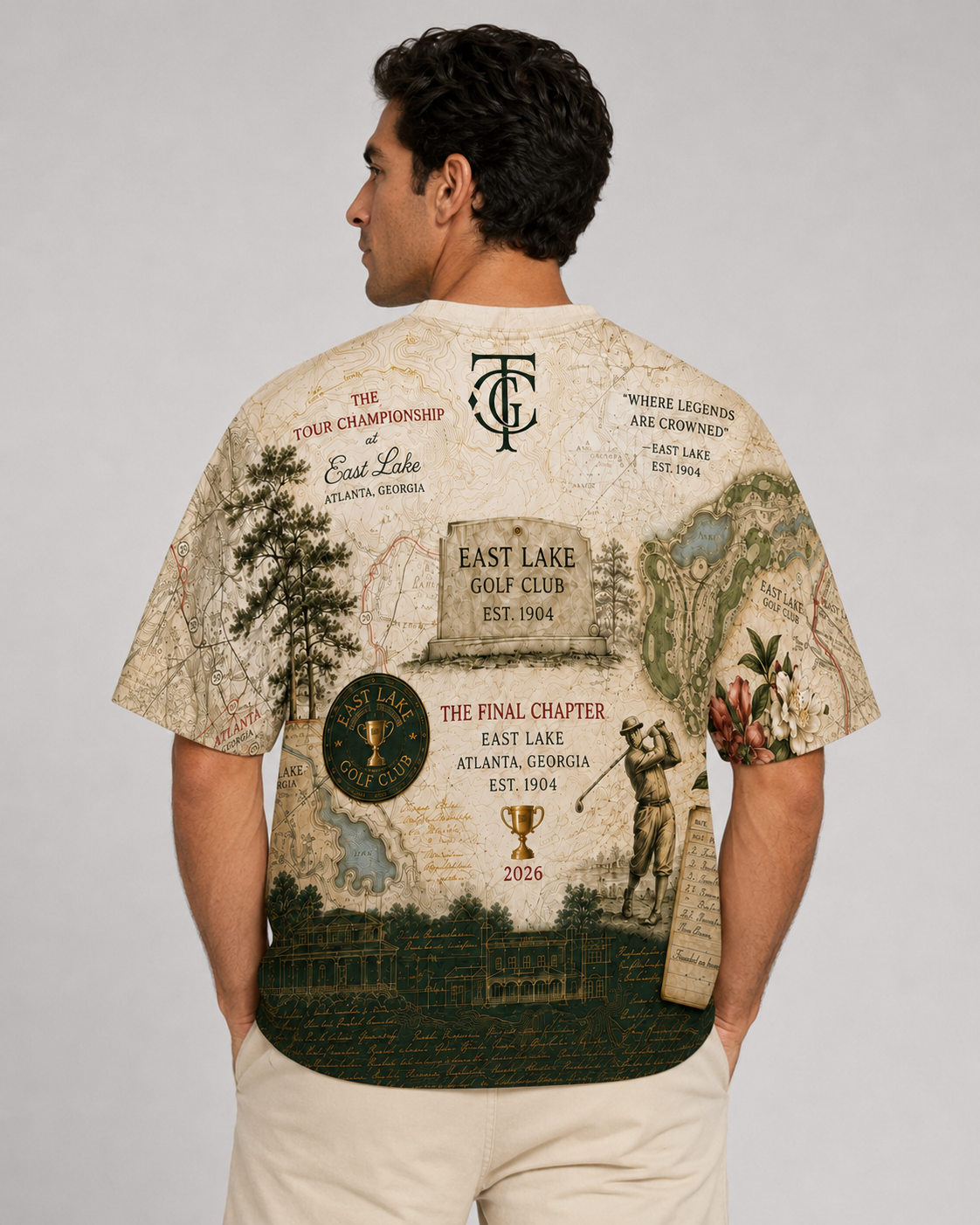 A man shown from the back wearing a vintage-style graphic t-shirt with a detailed East Lake Golf Club map print, paired with beige pants, against a neutral gray studio background.