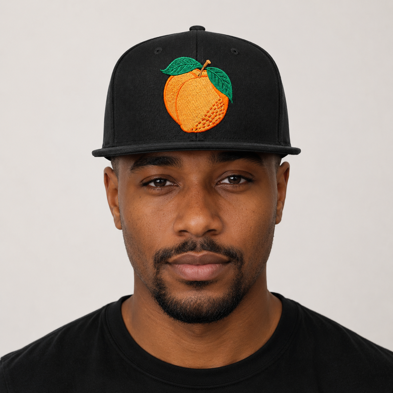Man wearing a black cap with an embroidered peach design on a plain background