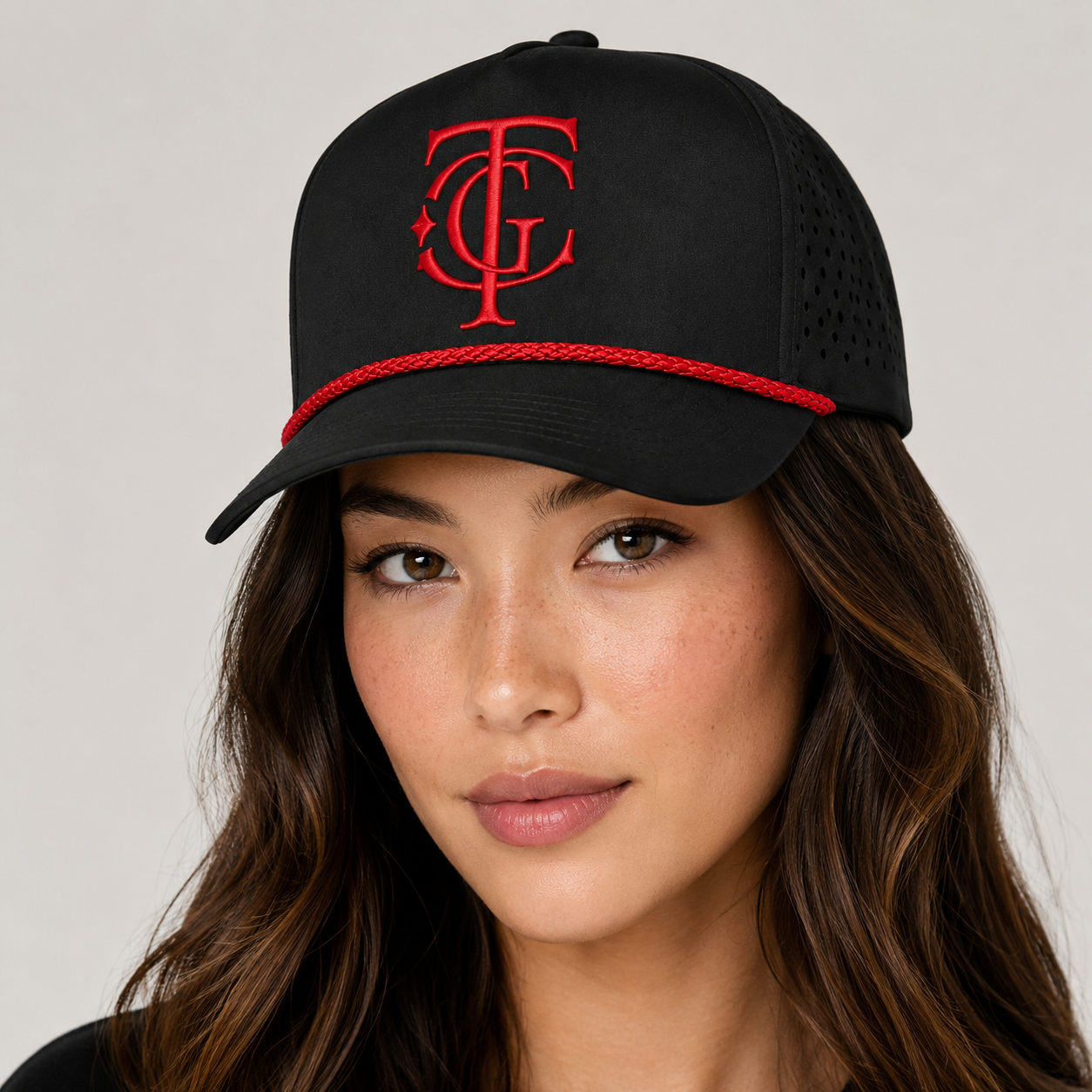 A woman wearing a black trucker-style cap with a red embroidered monogram logo on the front and a red braided rope detail around the brim, photographed against a light gray studio background.