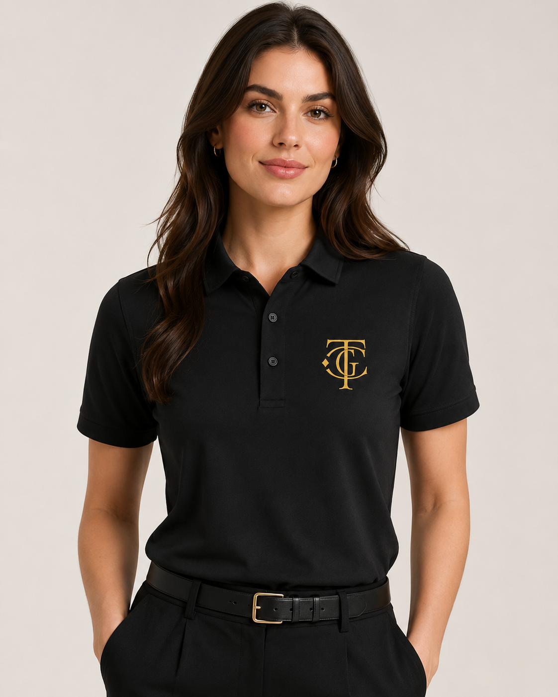A woman wearing a black short-sleeve polo shirt with a gold embroidered monogram logo on the left chest, paired with black trousers and a black belt, against a light beige studio background.