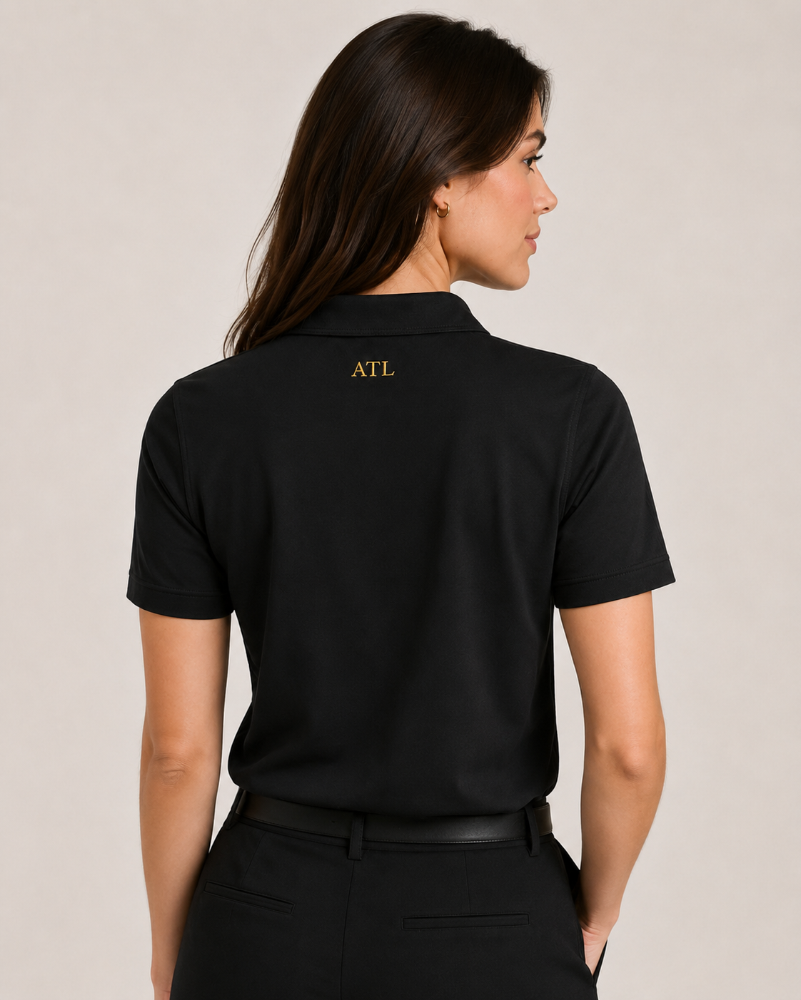 A woman shown from the back wearing a black short-sleeve polo shirt with 'ATL' embroidered in gold on the upper back, paired with black pants against a plain beige studio background.