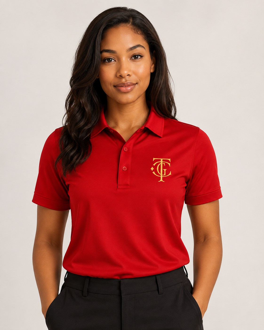 A woman wearing a bright red short-sleeve polo shirt with a gold embroidered monogram logo on the left chest, paired with black trousers, against a neutral light gray background.