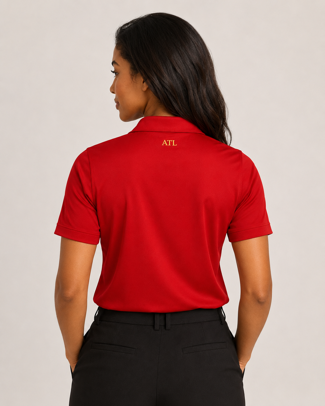 A woman viewed from the back wearing a bright red short-sleeve polo shirt with a small gold 'ATL' logo on the collar, paired with black pants, against a plain light gray studio background.