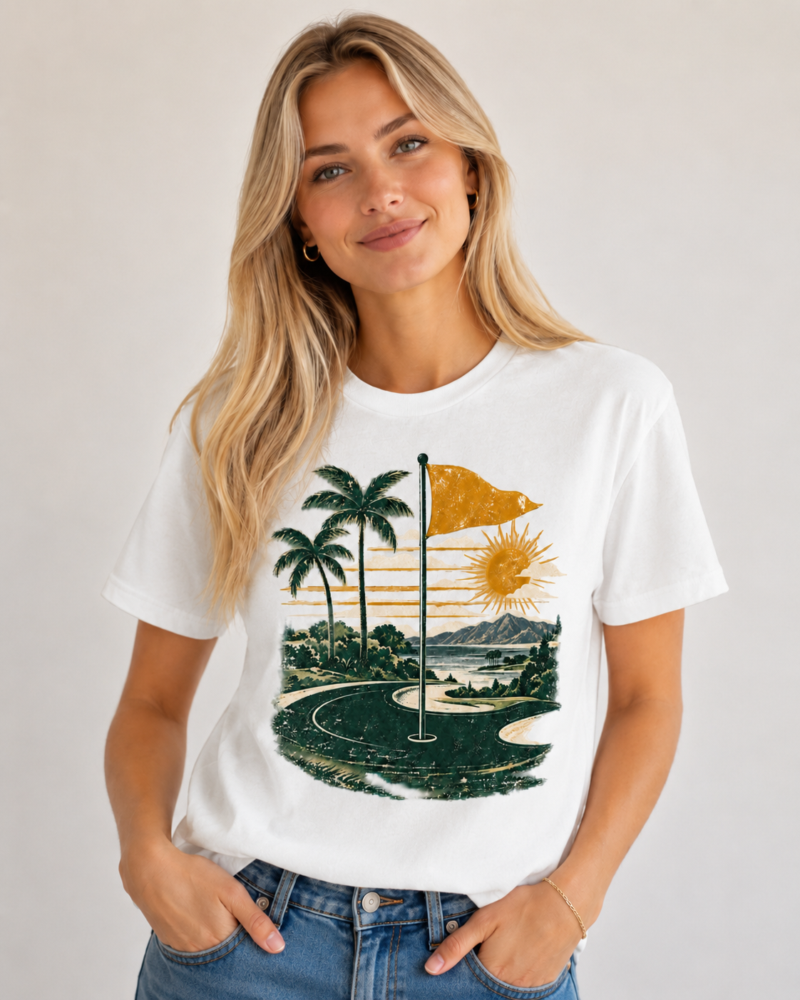 A woman wearing a white graphic t-shirt with a retro golf course sunset print, paired with blue jeans, against a neutral light gray background.