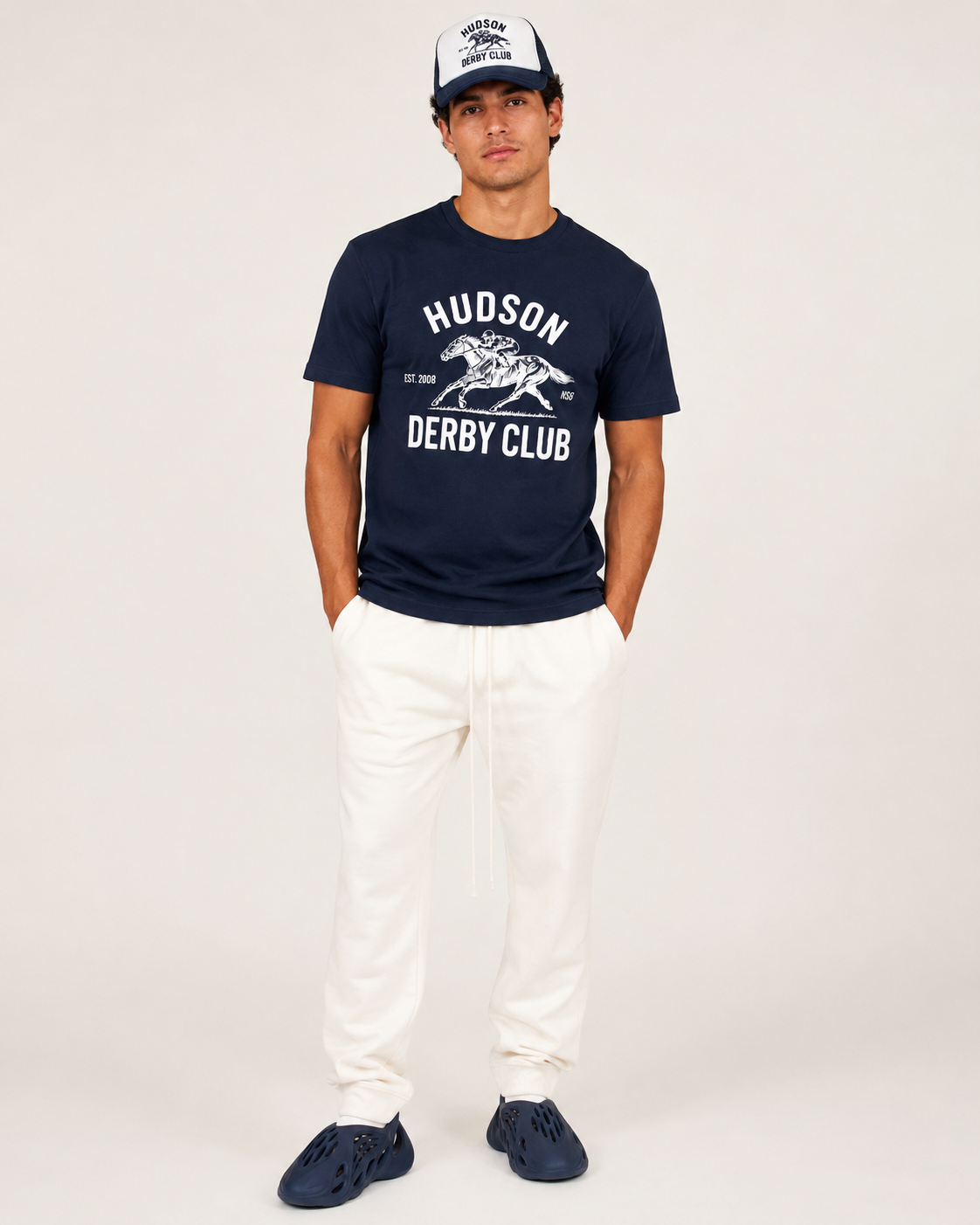 Man wearing a navy 'Hudson Derby Club' t-shirt and white pants on a white background