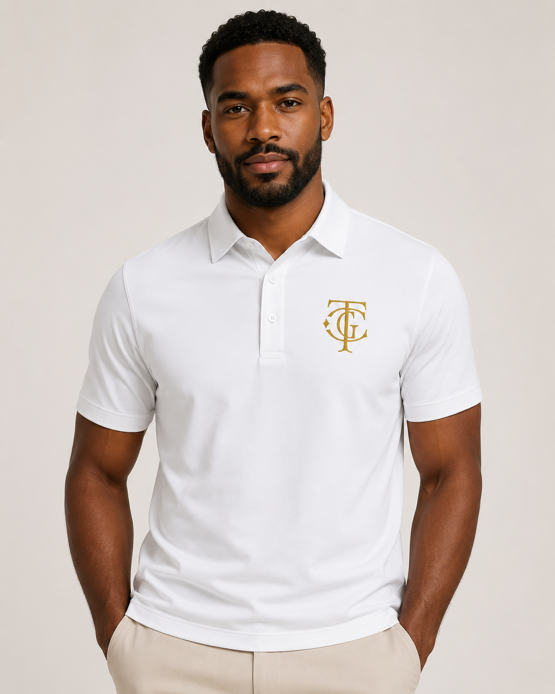 A man wearing a white short-sleeve polo shirt with a gold monogram logo on the left chest, paired with beige trousers, against a light beige studio background.