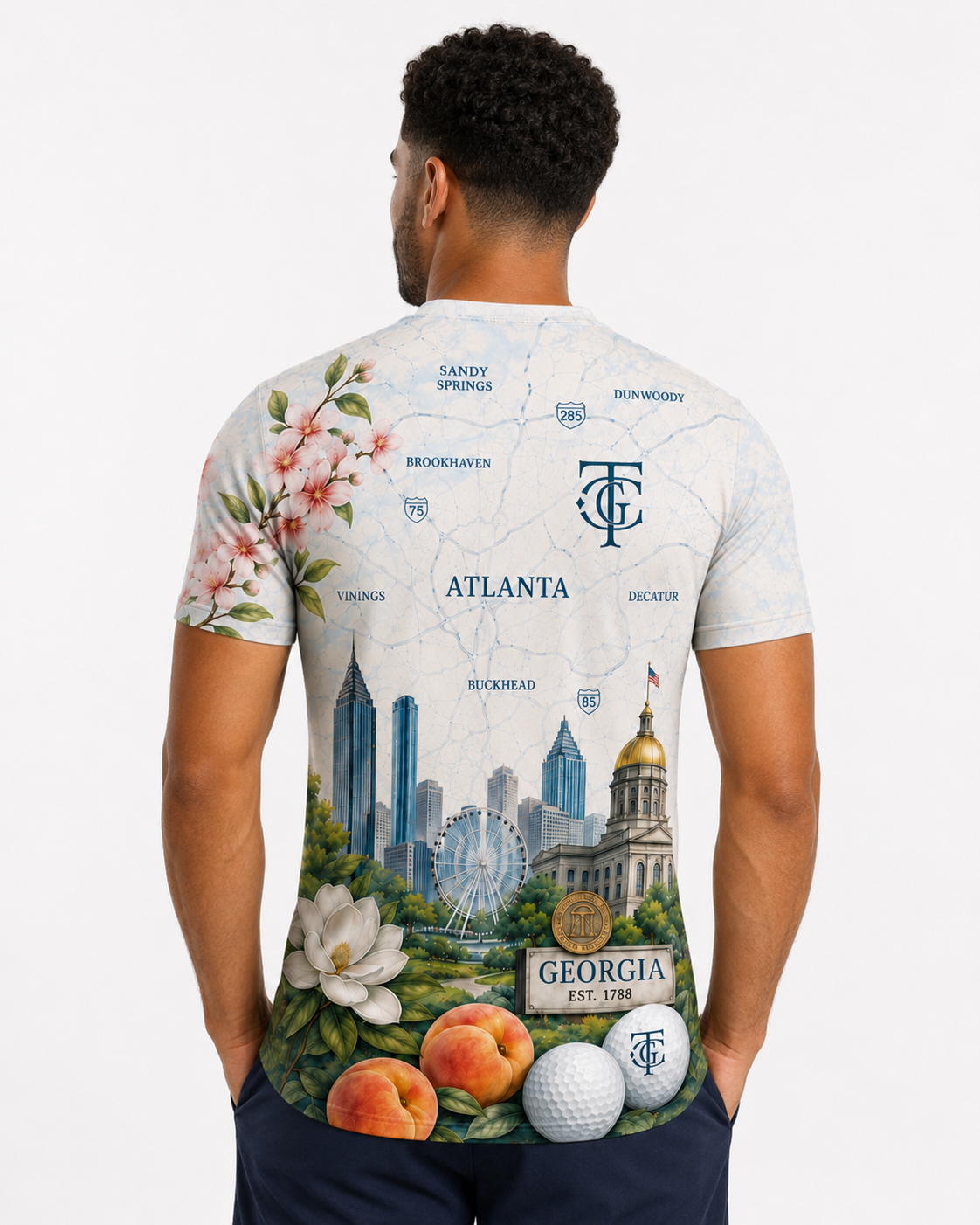 A man viewed from behind wearing a short-sleeve graphic t-shirt with a detailed Atlanta map design, featuring the Georgia state seal, skyline, flowers, peaches, and golf balls.