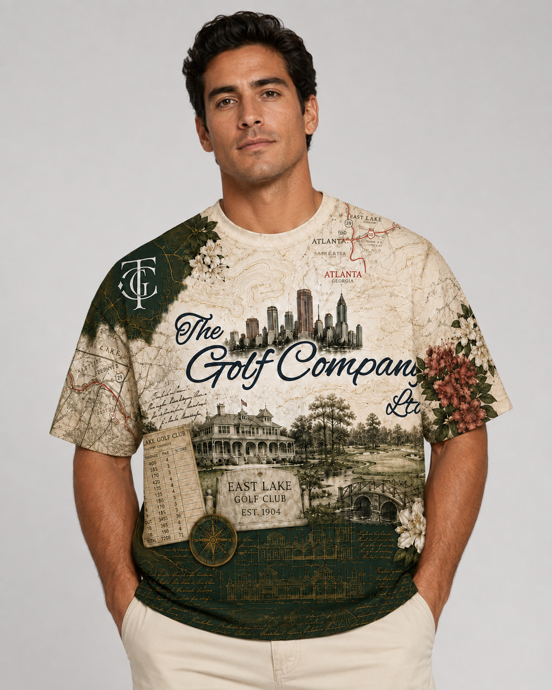 A man wearing a graphic t-shirt with a vintage-style golf course map print featuring 'The Golf Company Ltd.' and 'East Lake Golf Club' text, paired with off-white pants, against a neutral gray background.