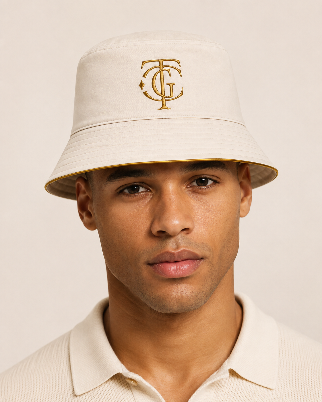 A man wearing a cream-colored bucket hat with a gold embroidered monogram logo on the front, paired with a matching cream polo shirt, set against a solid light beige background.