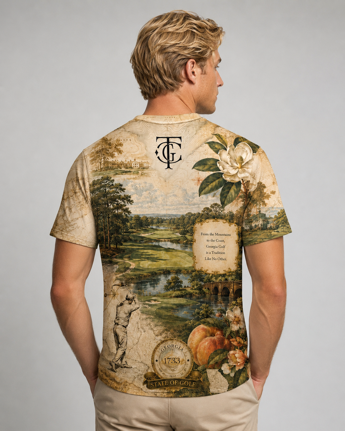 A man shown from the back wearing a vintage-style beige t-shirt with a detailed landscape print, featuring a golf course, flowers, and a monogram logo at the top.