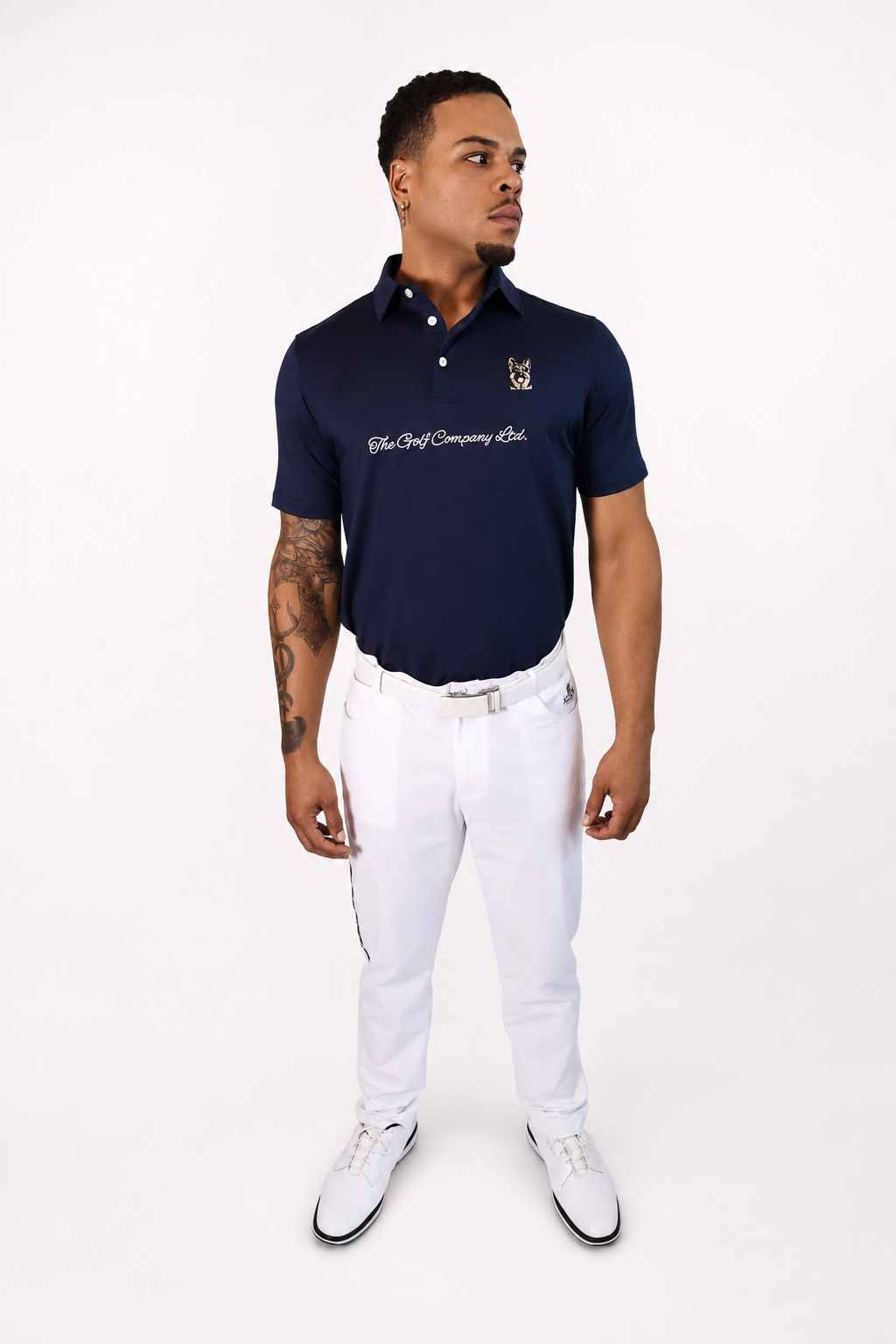 A man modeling a navy blue polo shirt with white embroidered text and a crest logo on the chest, paired with white trousers and white shoes, against a plain white studio background.