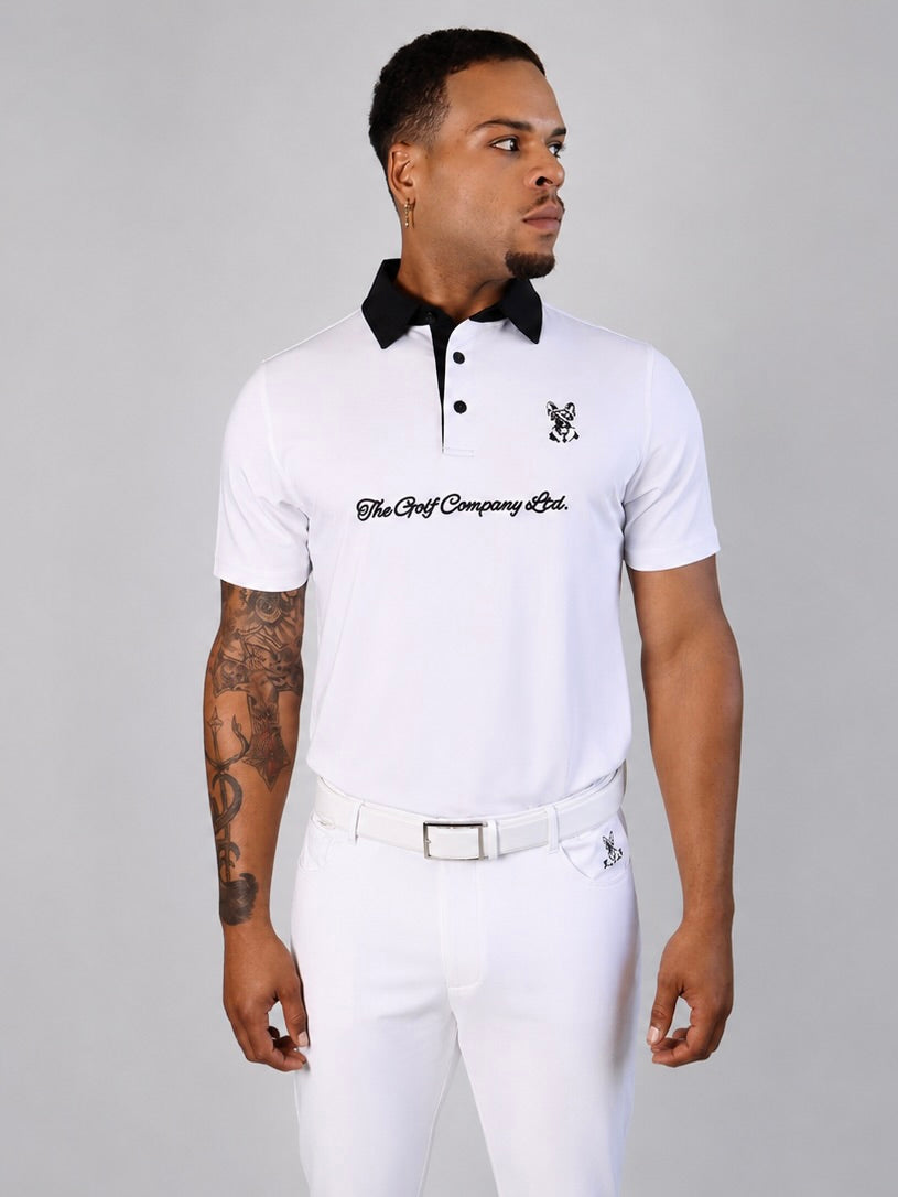 A man wearing a white short-sleeve polo shirt with a black collar, black buttons, and black script text across the chest, paired with white trousers, against a plain gray studio background.