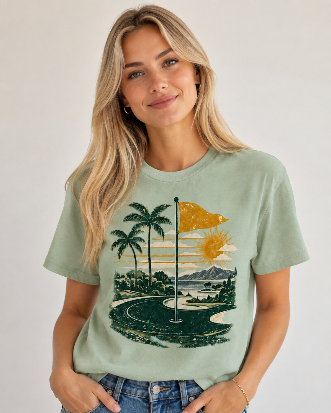 A woman wearing a sage green graphic t-shirt with a retro golf course design featuring palm trees, a flag, and a sunset, paired with blue jeans against a plain light gray background.